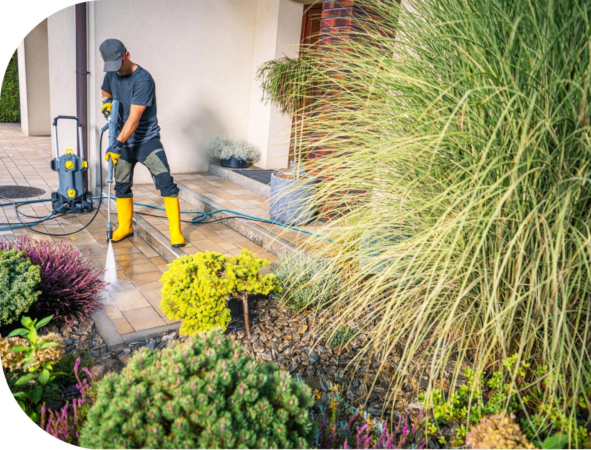 A person in yellow rubber boots and gloves uses a pressure washer to clean a tiled patio surrounded by plants and shrubs, demonstrating outdoor maintenance work.