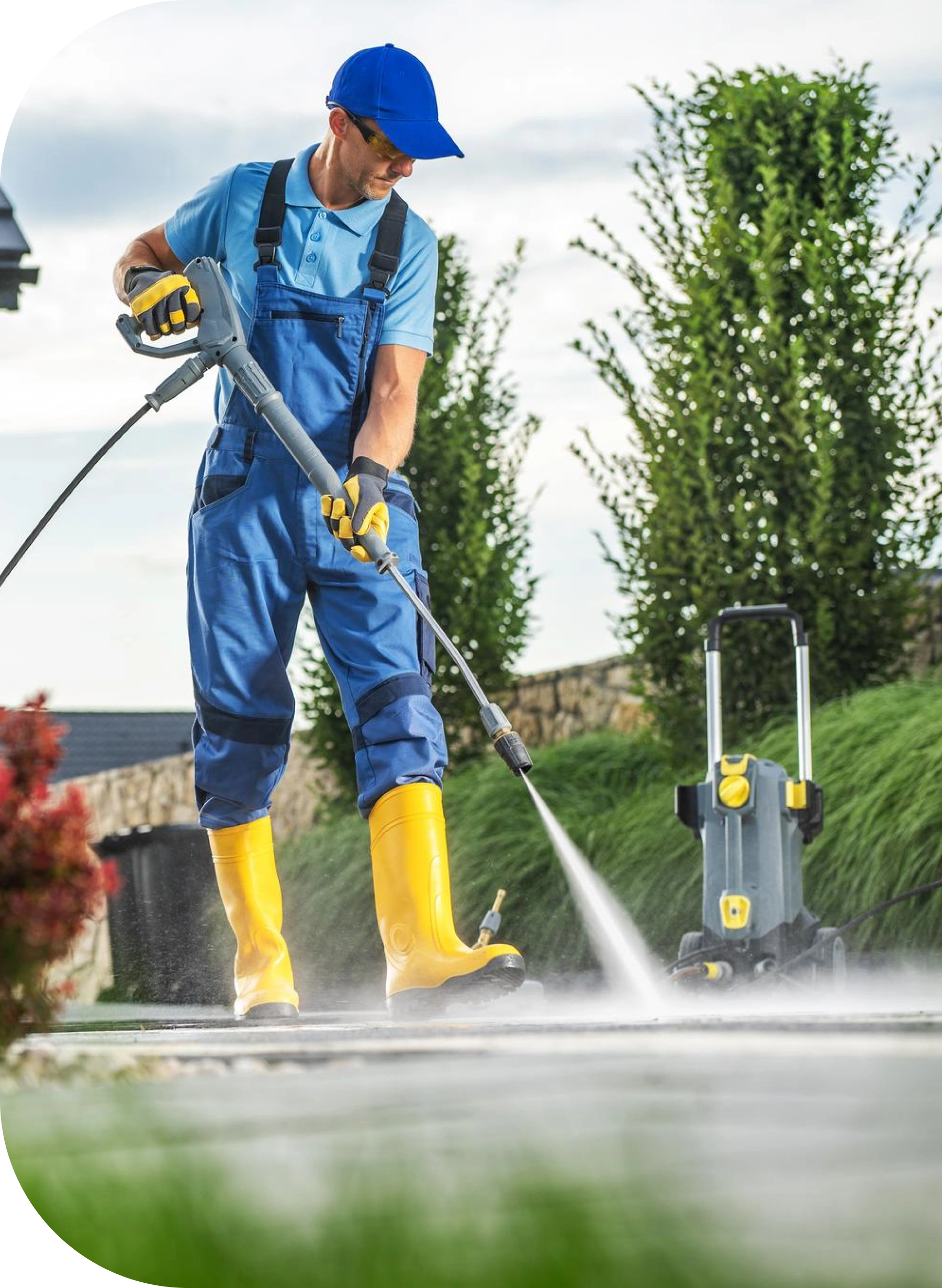 A person in blue overalls, yellow boots, gloves, and a cap uses a portable pressure washer to clean a paved outdoor surface, with green grass and tall trees in the background