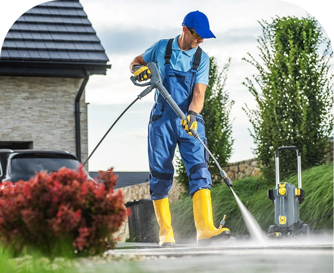 A person in blue overalls, yellow boots, gloves, and a cap uses a portable pressure washer to clean a paved outdoor surface, with green grass and tall trees in the background