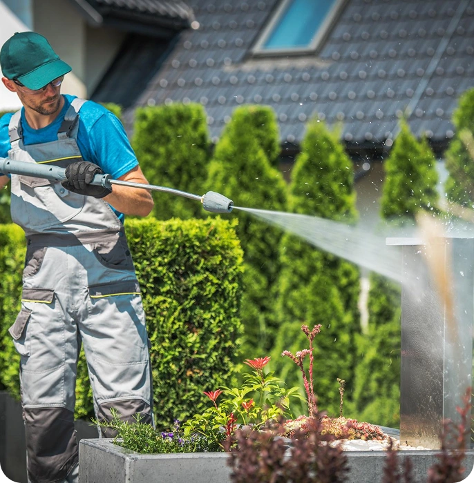 A person in gray overalls, teal shirt, gloves, and cap uses a pressure washer to clean an outdoor metallic or stone structure in a landscaped garden with hedges and a house in the background.