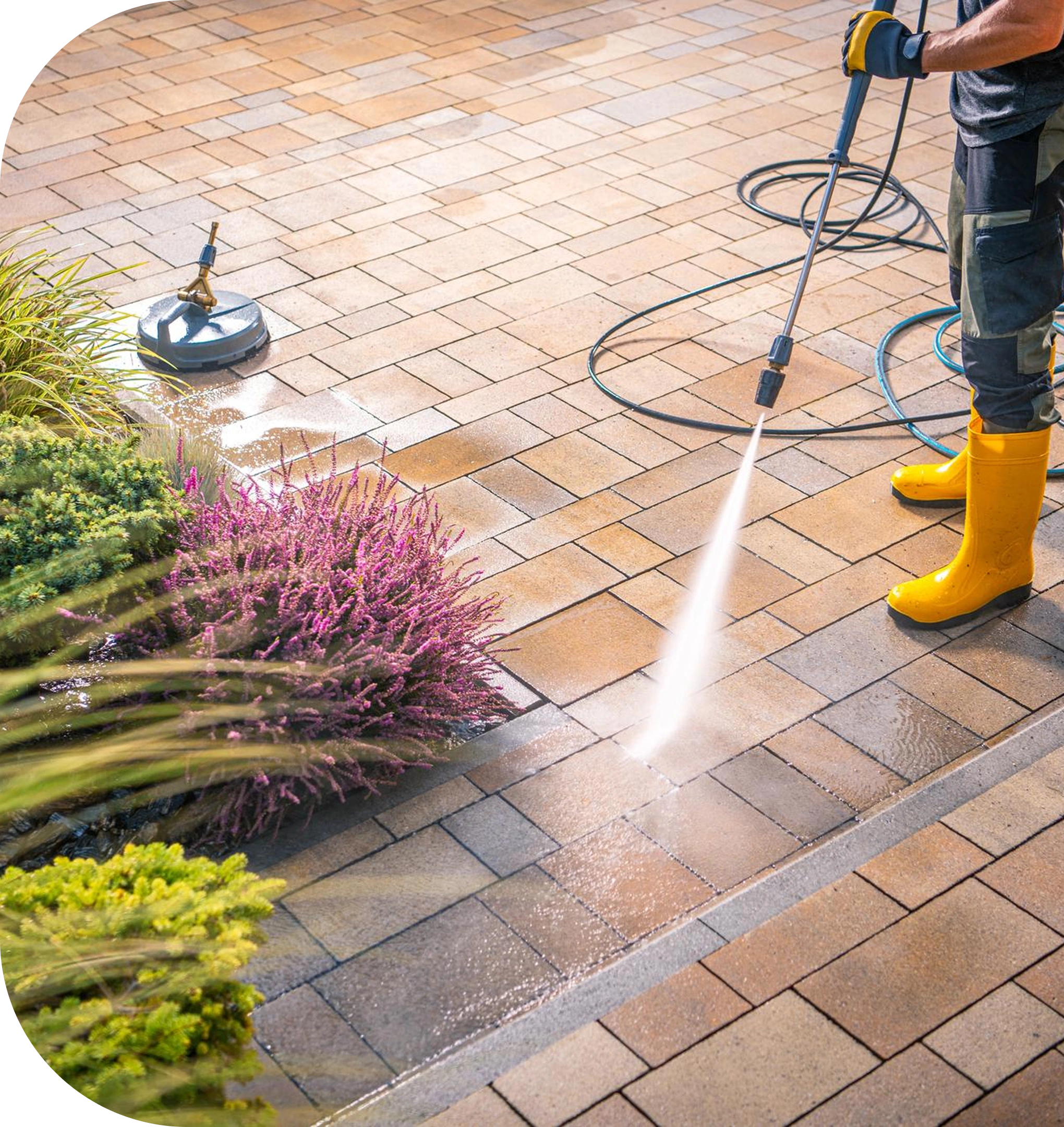A person pressure washing a stone patio, wearing yellow boots and gloves, with water visibly cleaning the rectangular tiles. A hose and surface cleaner attachment lie nearby, and plants border the edge of the paved area.