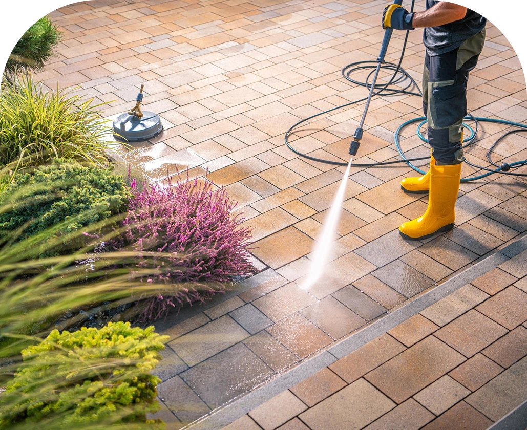 A person pressure washing a stone patio, wearing yellow boots and gloves, with water visibly cleaning the rectangular tiles. A hose and surface cleaner attachment lie nearby, and plants border the edge of the paved area.