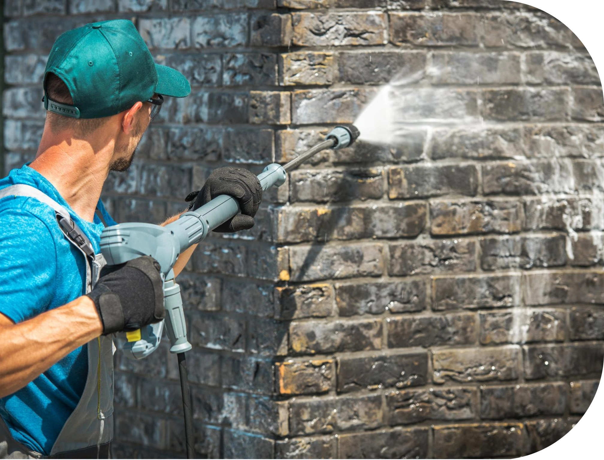A person pressure washing a brick wall, wearing a teal cap, blue shirt, and black gloves, as a strong jet of water cleans dirt from the dark bricks.