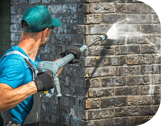 A person pressure washing a brick wall, wearing a teal cap, blue shirt, and black gloves, as a strong jet of water cleans dirt from the dark bricks.
